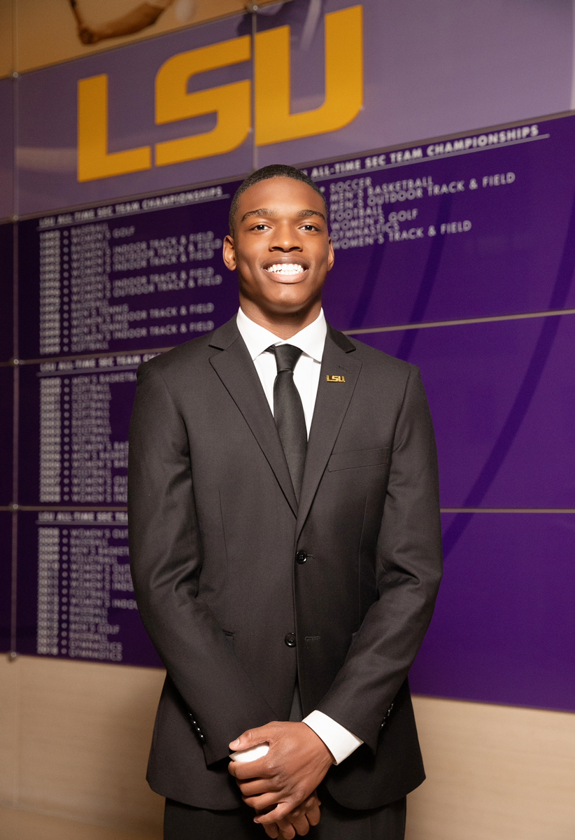 Zyan Williams wears a dark formal suit. He smiles as he stands in front of a wall wiht the names of LSU champions.