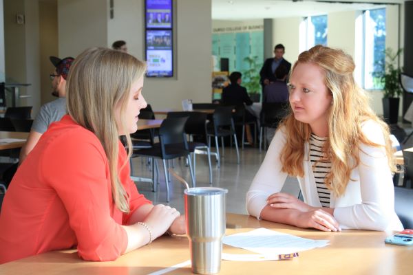 Woman in an orange shirt sits across a table from a female college student. 